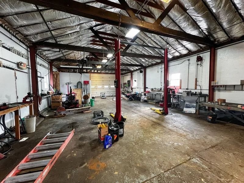 Empty industrial workshop with metal roof, red support columns, tools, ladders, and equipment scattered on the floor