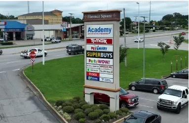 Shopping plaza sign listing stores by a road, with parked cars and green landscaping.