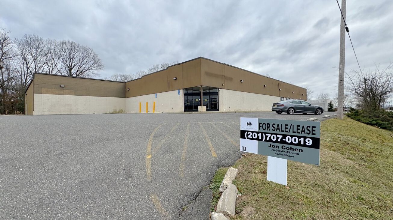 Empty gravel lot with a beige warehouse and a for-sale sign in front