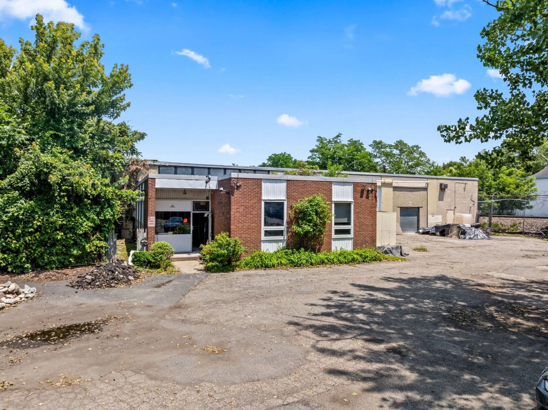 Abandoned low brick building with boarded windows, gravel lot, and trees under a blue sky