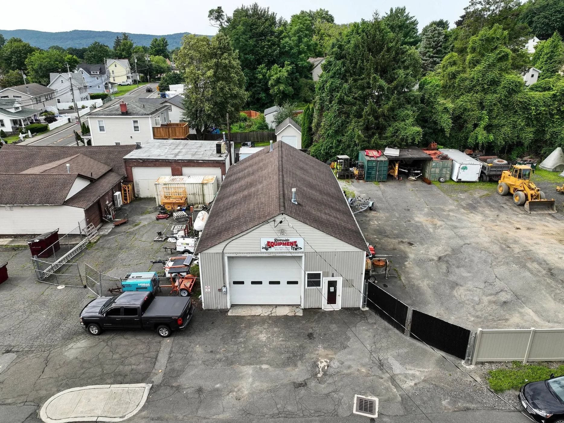 Aerial view of an auto repair shop with trucks, equipment, and a fenced dirt lot beside trees and houses.