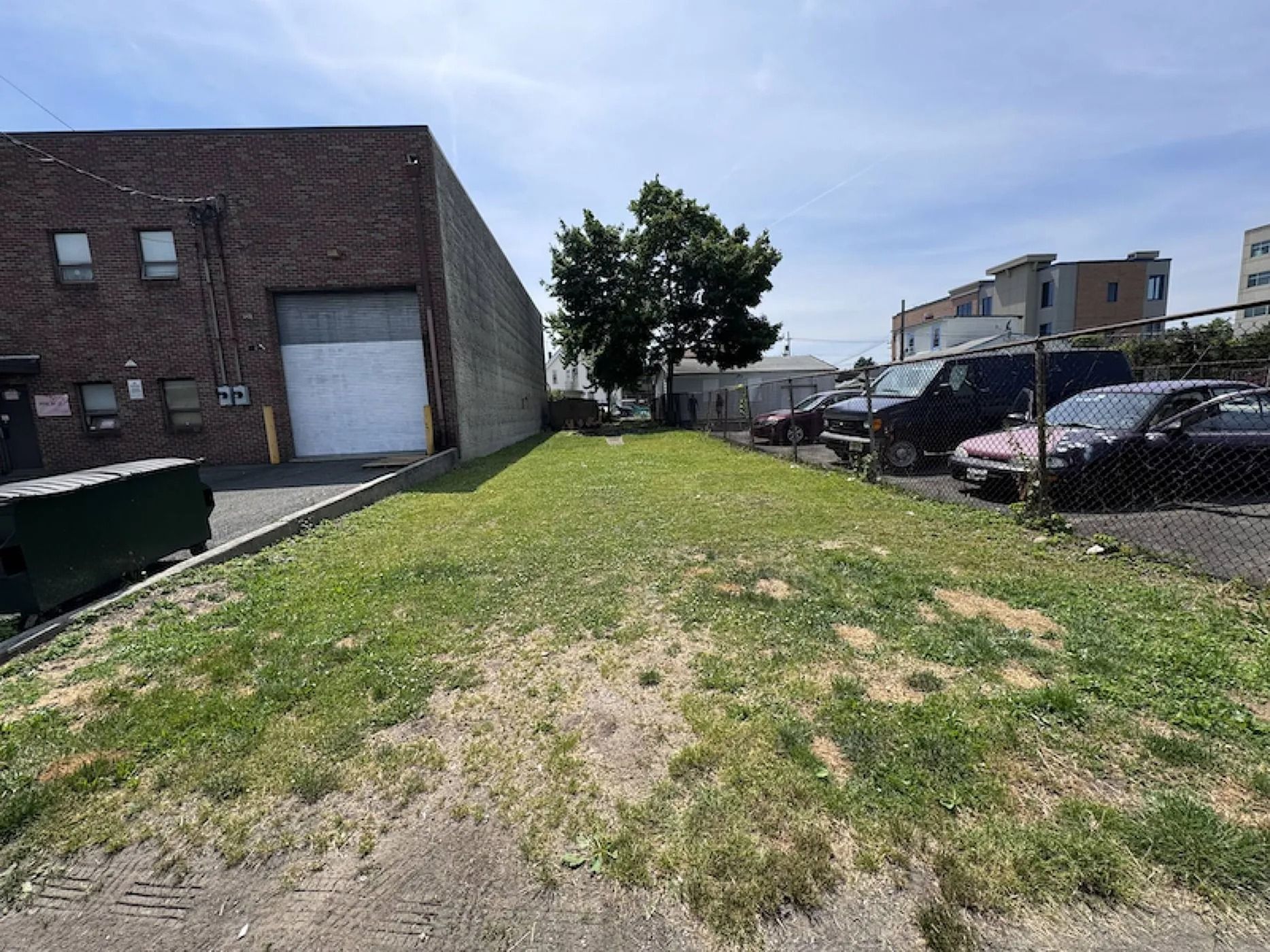 Narrow grassy strip beside a brick building, with parked cars and houses under a partly cloudy sky.