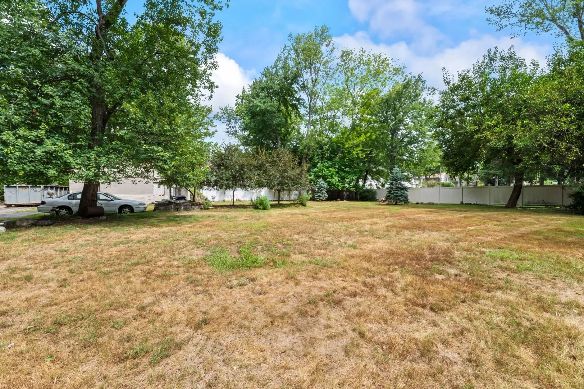 Wide grassy backyard with mature green trees, a white fence, and blue sky with clouds