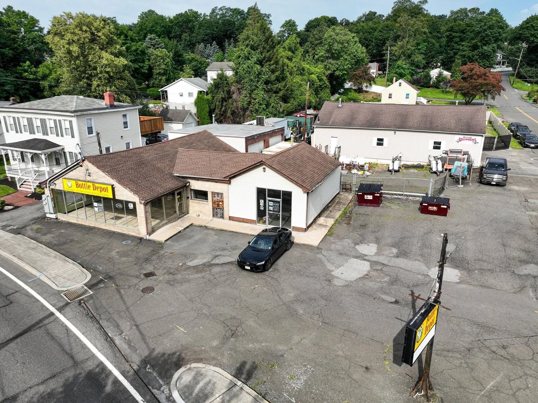Aerial view of a small commercial building and parking lot beside a road, with trees and houses behind it