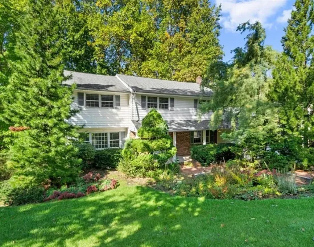 White two-story house with a brick entry, surrounded by lush green trees and a manicured lawn.