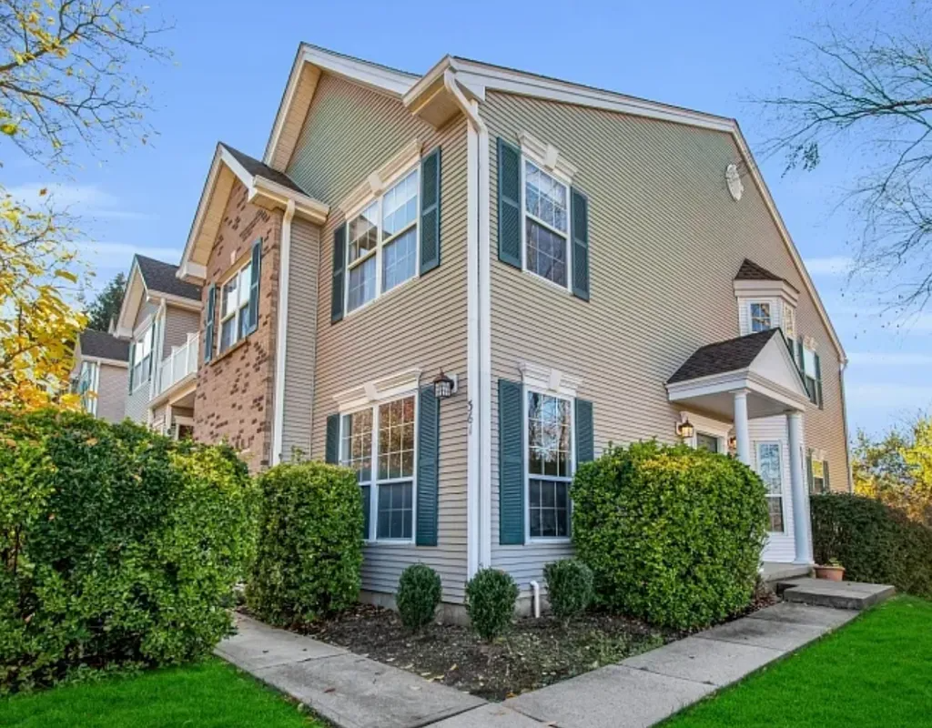 Two-story beige townhouse with white trim, brick accents, and green hedges along a sidewalk on a sunny day
