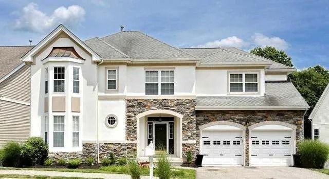 Large two-story beige house with white trim, stone accents, and a three-car garage under a blue sky