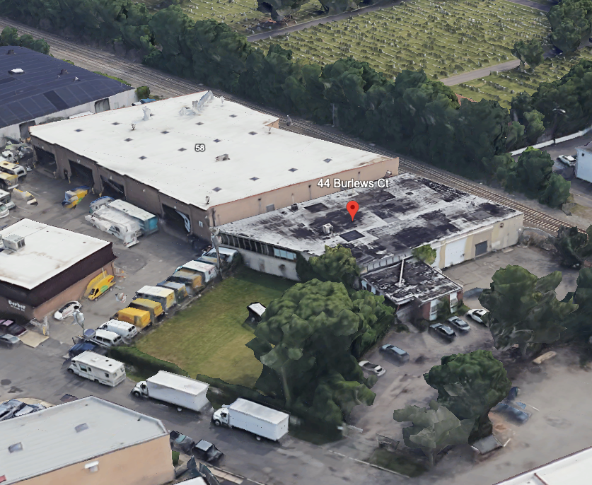 Aerial view of an industrial warehouse lot with parked trucks, trees, and a red location pin on the roof.