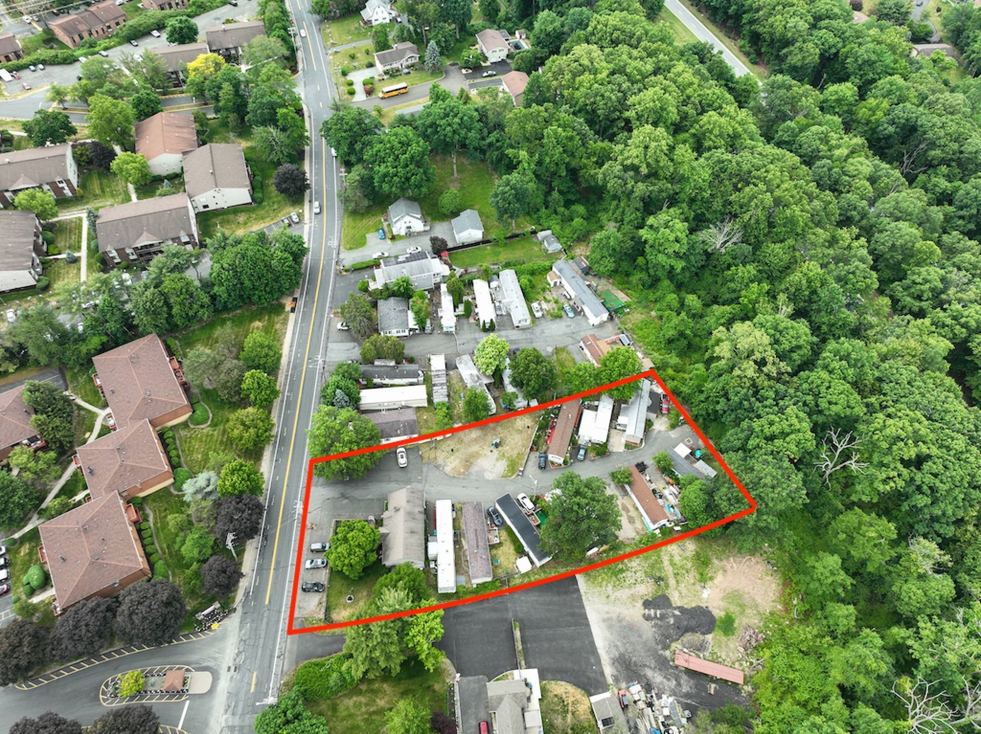 Aerial view of a residential area with houses, roads, and a red outlined property beside dense green trees