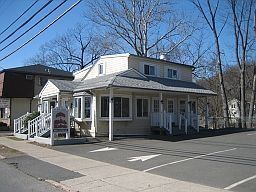 Small white building with porch and stairs beside a parking lot on a sunny street corner