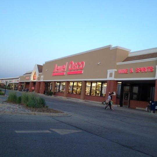 Strip mall storefronts at dusk, with a large grocery sign and a person walking by the entrance.