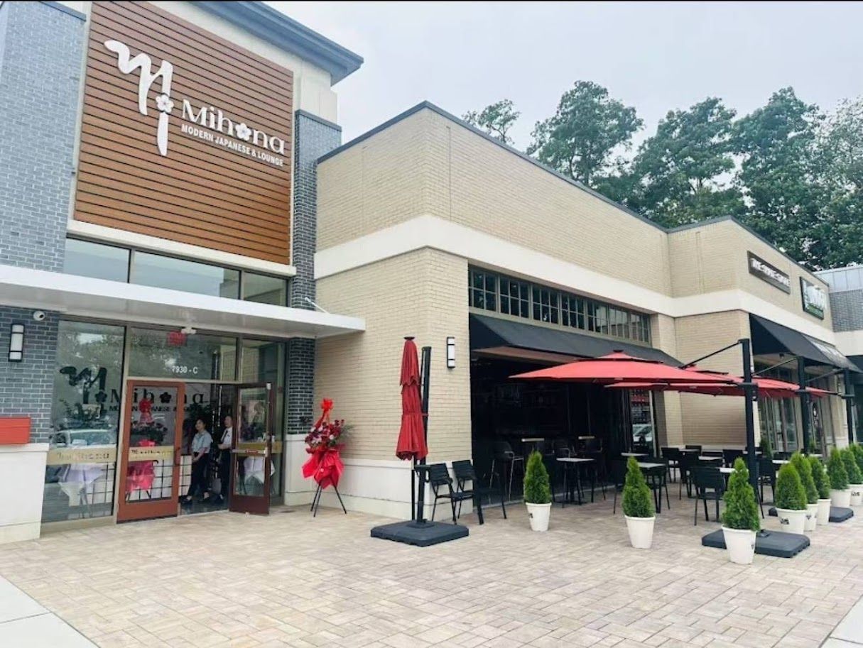 Outdoor shopping plaza with storefronts, red umbrellas, and a person in a red outfit near the entrance