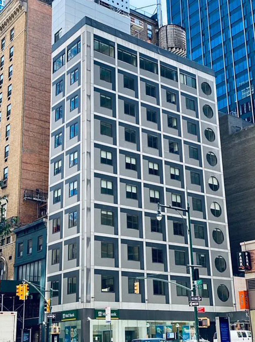 Modern gray office building with many windows on a city street, framed by taller buildings