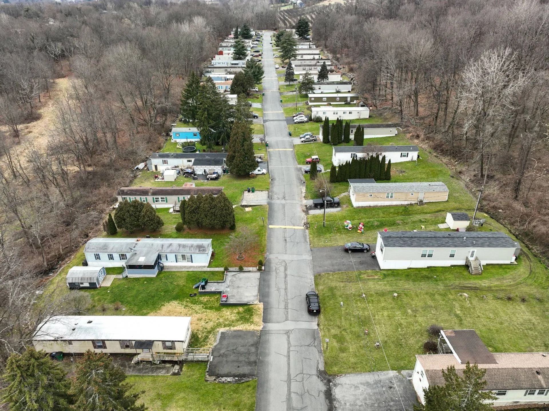 Aerial view of a suburban mobile home park with a straight road and rows of homes beside wooded areas