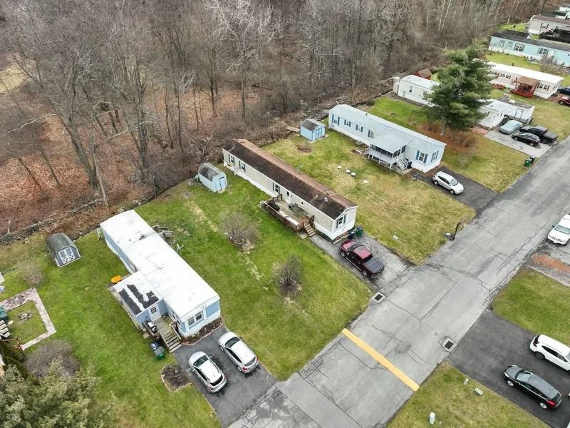Aerial view of a mobile home park with several trailers, parked cars, and a road beside leafless woods.