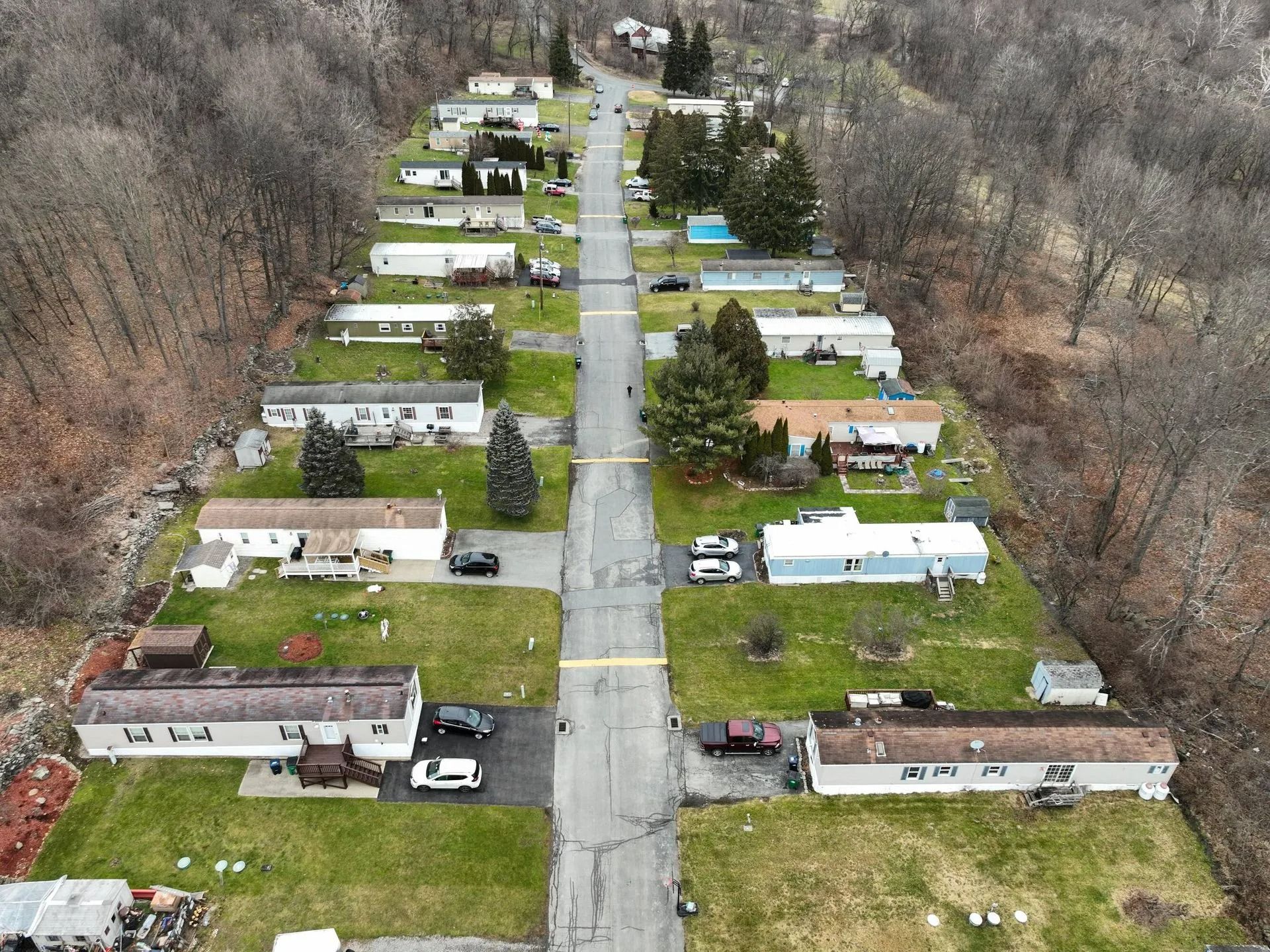 Aerial view of a mobile home park along a narrow road, with trailers on both sides beside wooded hills.
