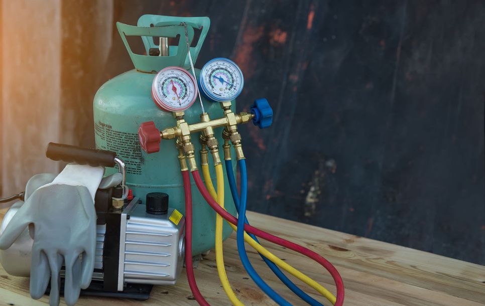 Refrigerant tank with pressure gauges and colored hoses on a wooden bench, beside gloves and tools