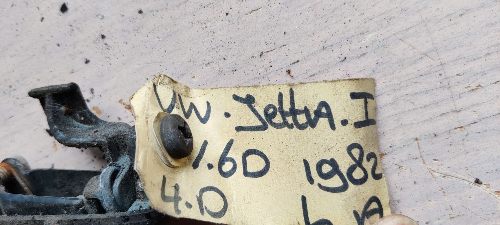 Een zwarte autodeurhendel met een klein papieren labeltje eraan, van de zijkant gezien tegen een witte achtergrond.