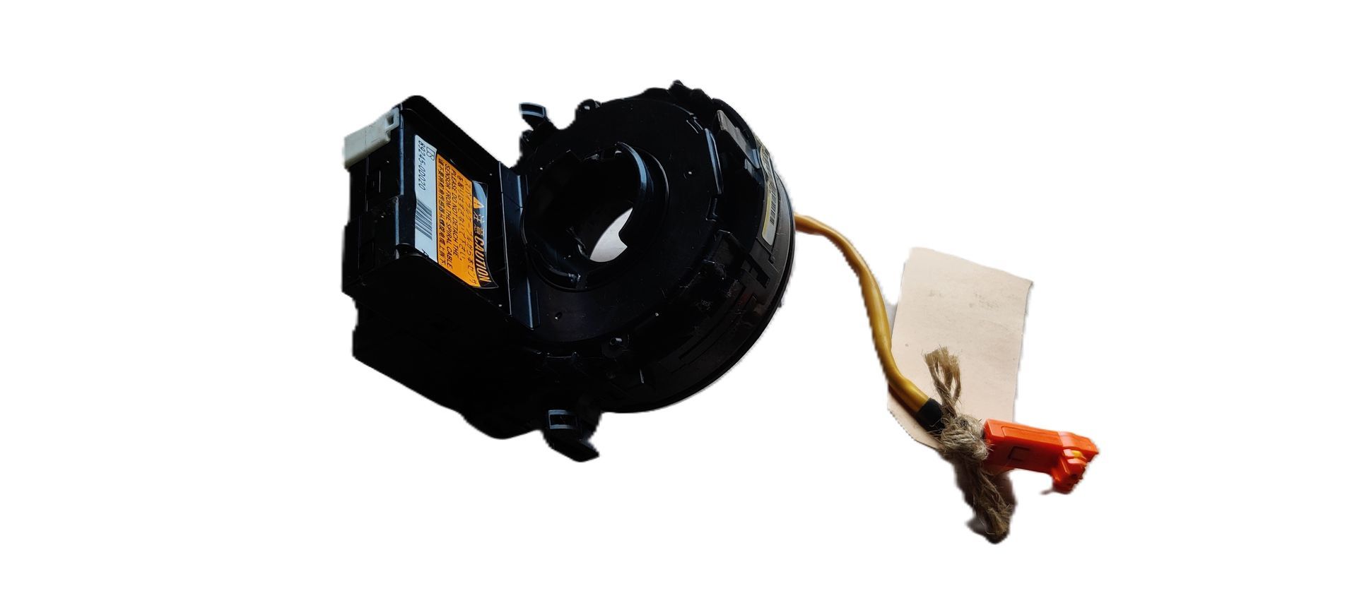 Een zwart auto-onderdeel, een klokveer, met een oranje bedradingsconnector en een klein wit labeltje.