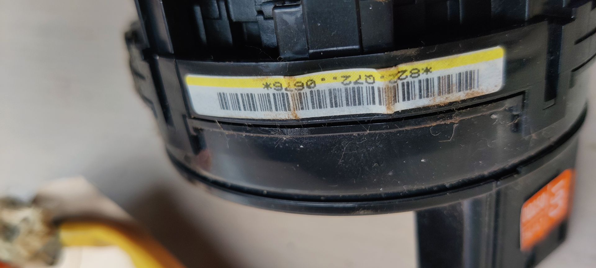 Een zwart auto-onderdeel, een klokveer, met een oranje bedradingsconnector en een klein wit labeltje.