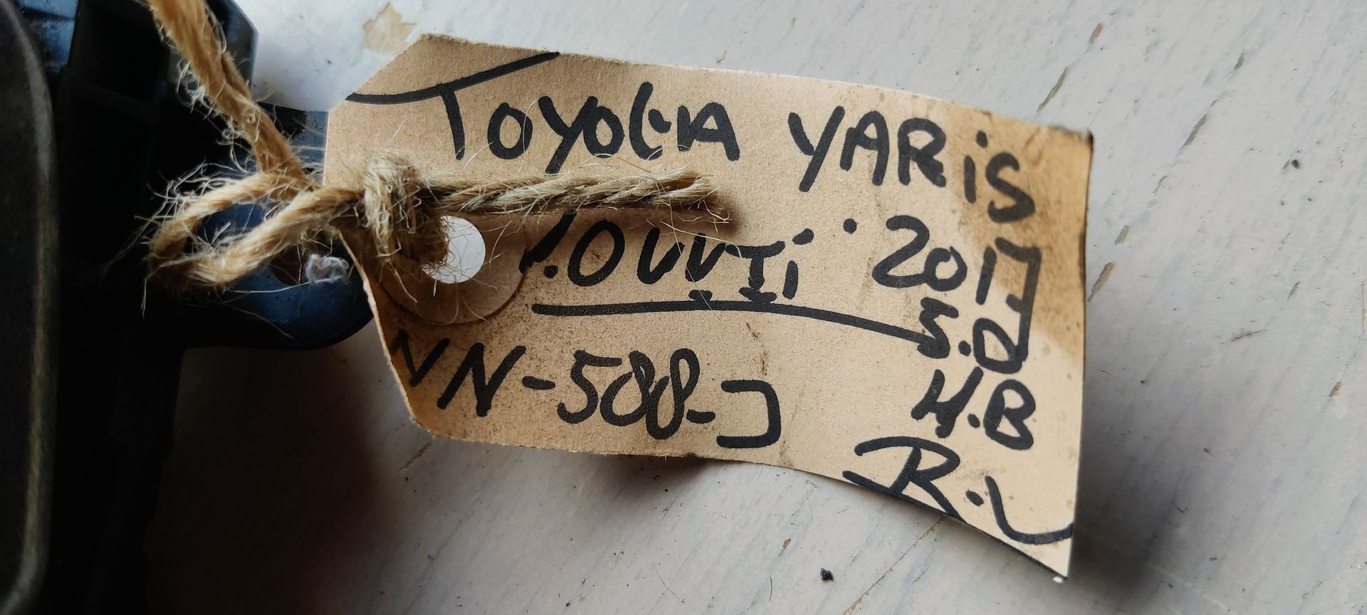 Een zwarte Toyota-deurhendel aan de buitenkant met een klein papieren labeltje eraan, afgebeeld tegen een witte achtergrond.