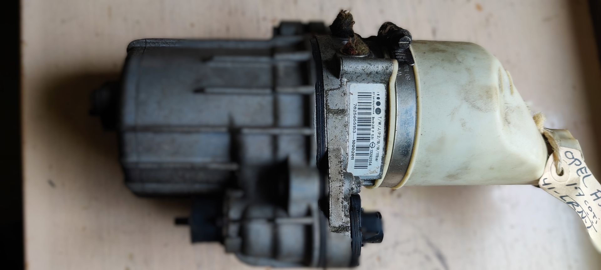 Een grijze elektrische stuurbekrachtigingsmotor voor auto's met een doorschijnend plastic reservoir en een barcode-sticker.