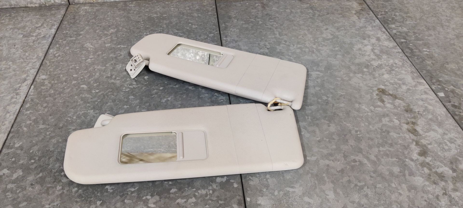 Twee beige zonnekleppen voor auto's met ingebouwde spiegels, liggend op een grijze tegelvloer.