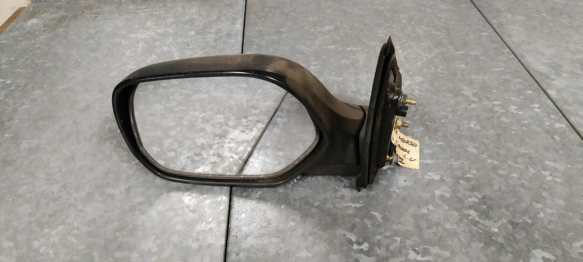 Een zwarte zijspiegel voor auto's met montagebeugel en bevestigingsmateriaal, rustend op een grijs metalen oppervlak.