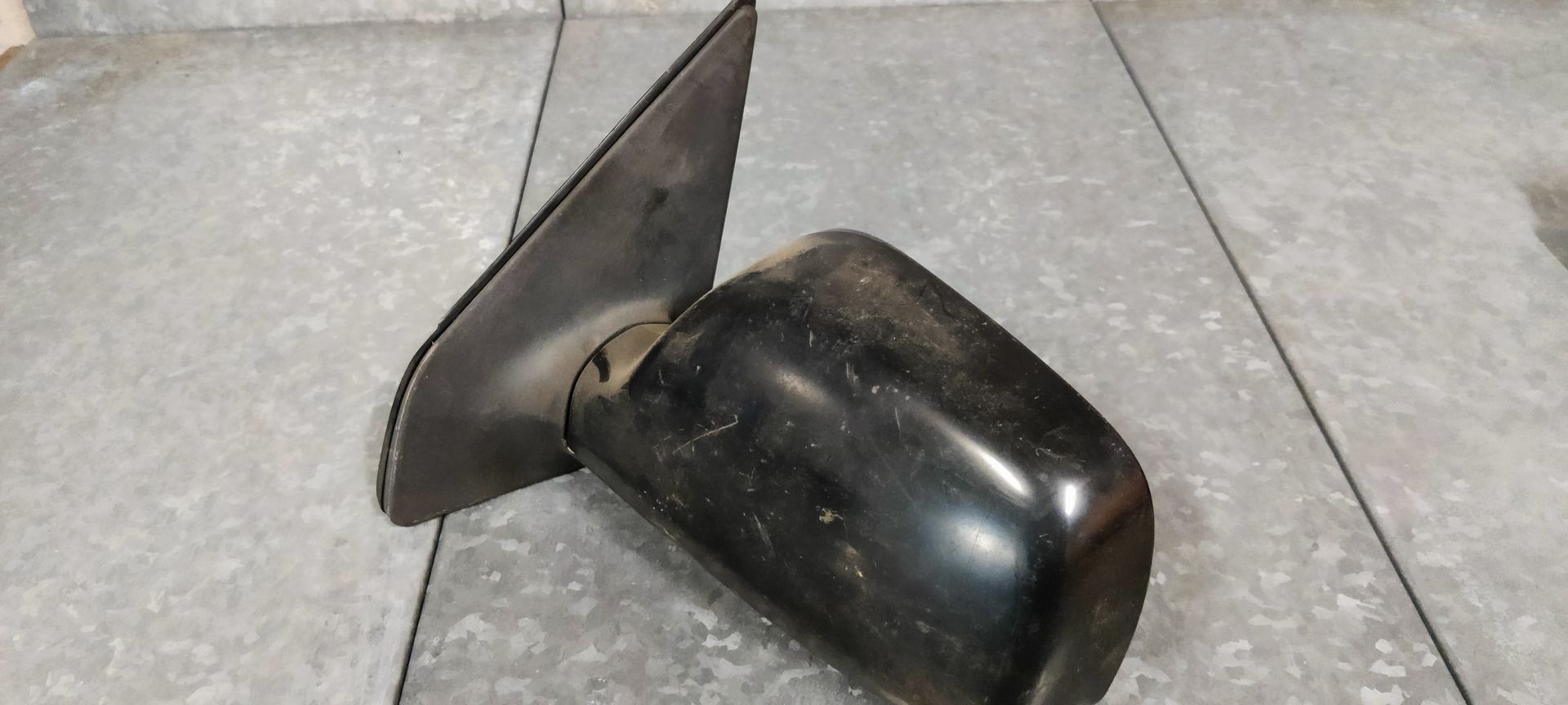 Een zwarte zijspiegel voor auto's met montagebeugel en bevestigingsmateriaal, rustend op een grijs metalen oppervlak.