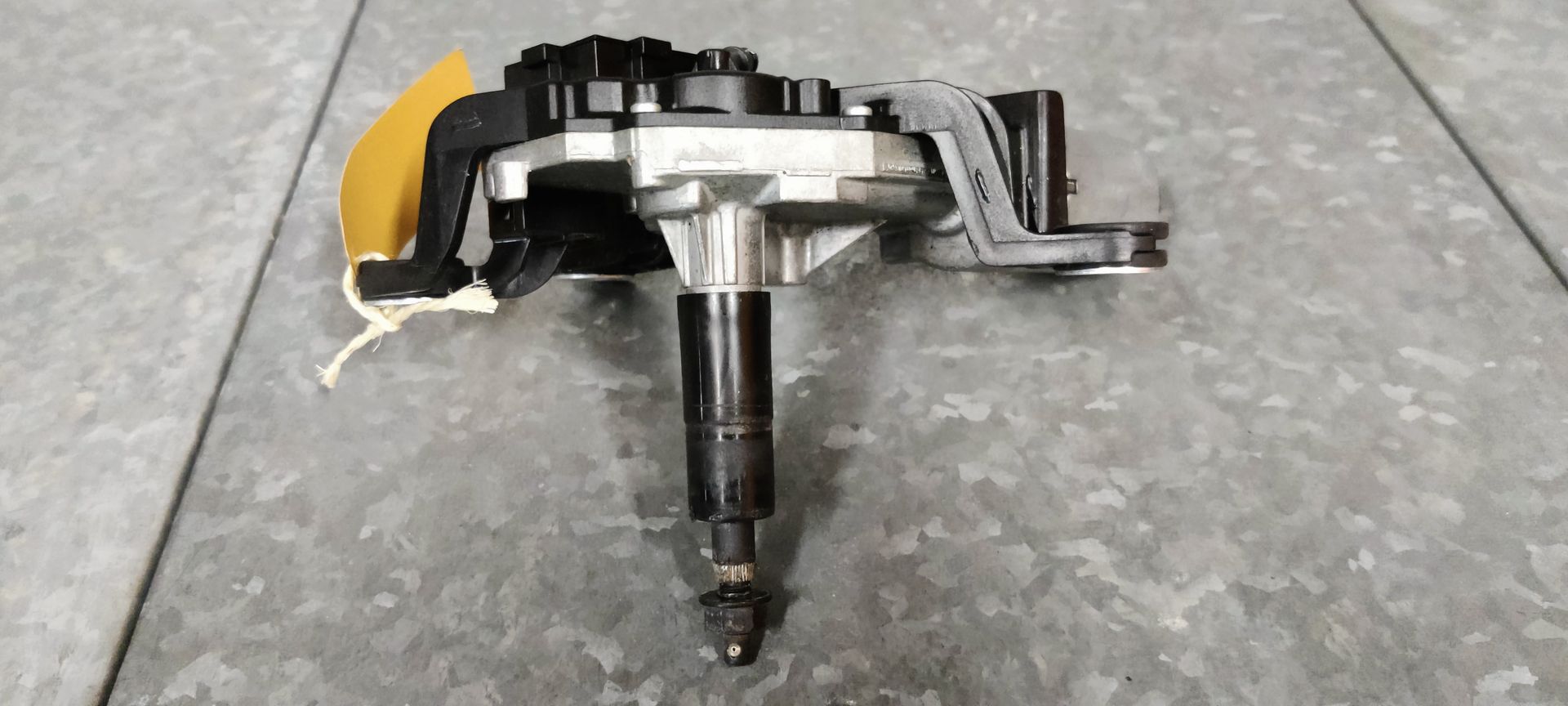 Een zwart-zilveren ruitenwissermotor met een uitstekende aandrijfas, rustend op een grijs betegeld oppervlak.