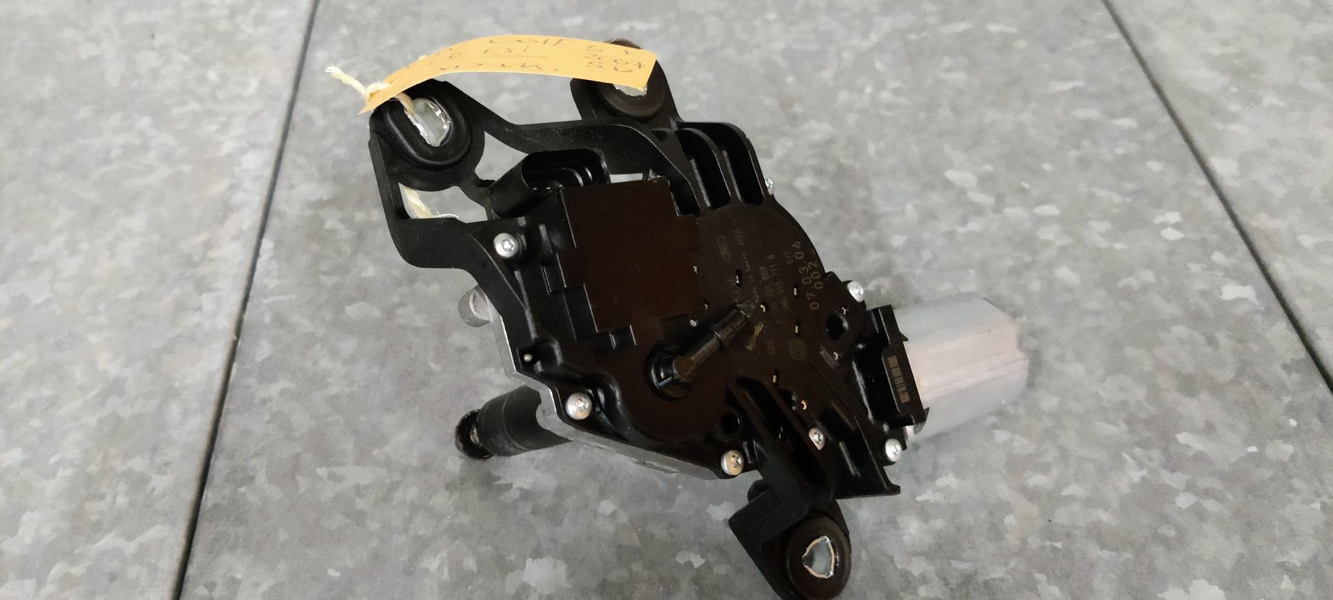 Een zwart-zilveren ruitenwissermotor met een uitstekende aandrijfas, rustend op een grijs betegeld oppervlak.