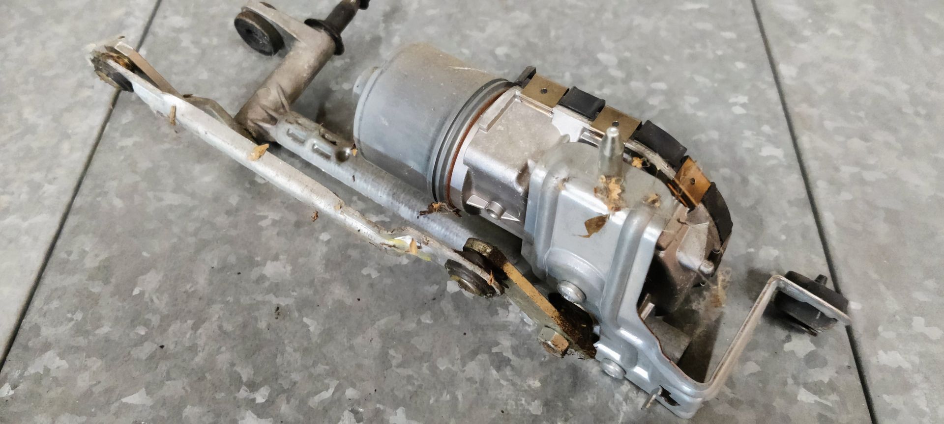 Een gebruikte ruitenwissermotor met bijbehorende koppeling op een grijze werkplaatsvloer.