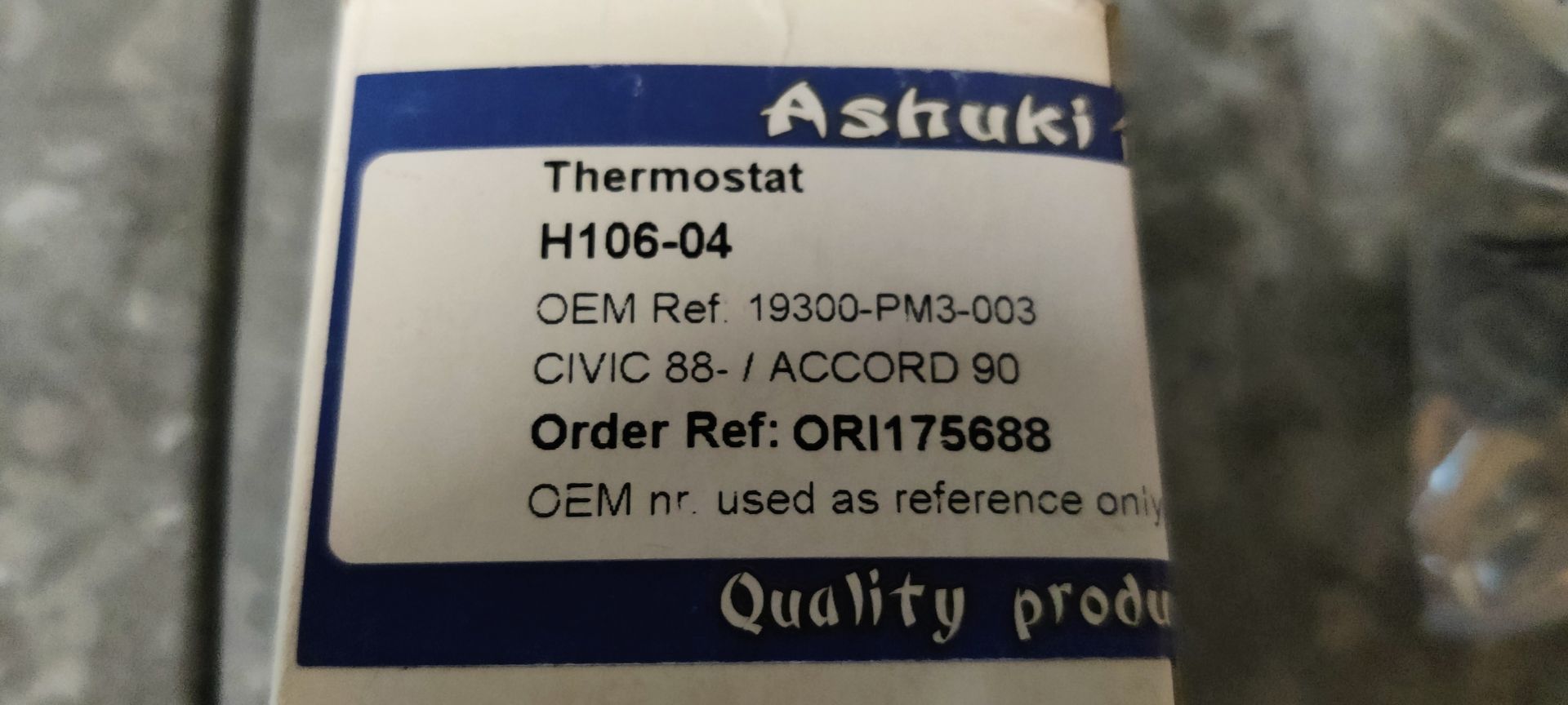 Een autothermostaat in een plastic zak naast de kartonnen doos op een grijze ondergrond.