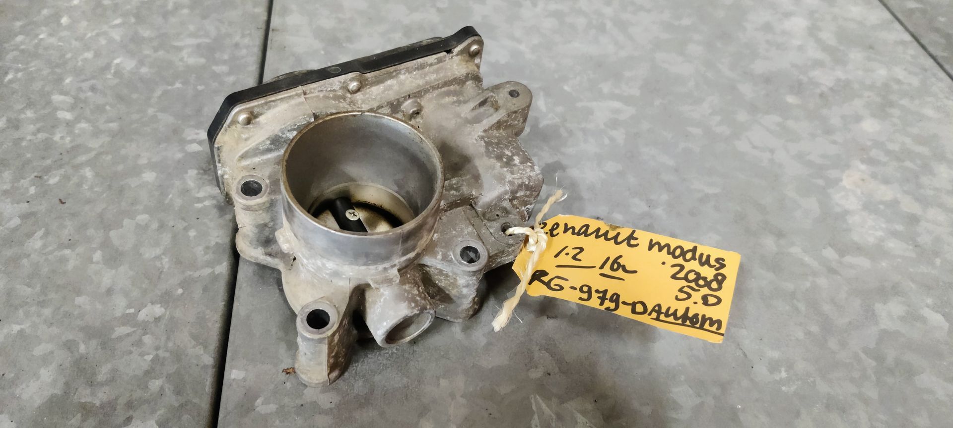 Een gebruikt metalen gasklephuis voor auto's, met een geel identificatielabel eraan, ligt op een grijze betonnen ondergrond.