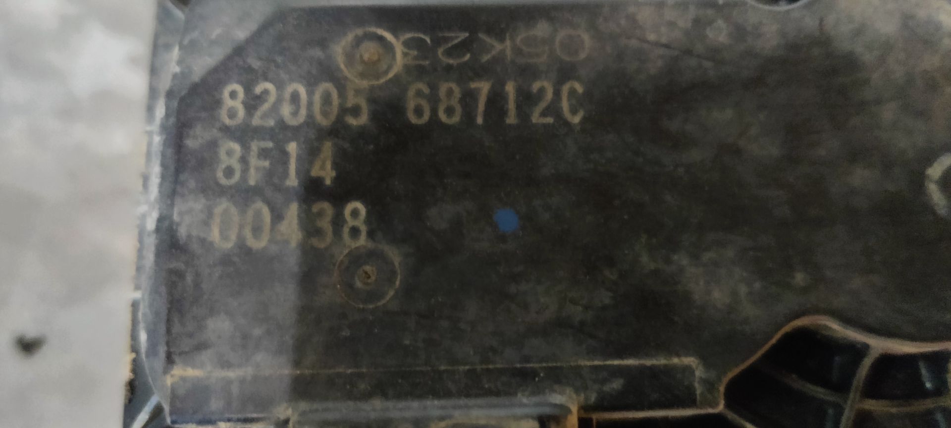 Een gebruikt metalen gasklephuis voor auto's, met een geel identificatielabel eraan, ligt op een grijze betonnen ondergrond.