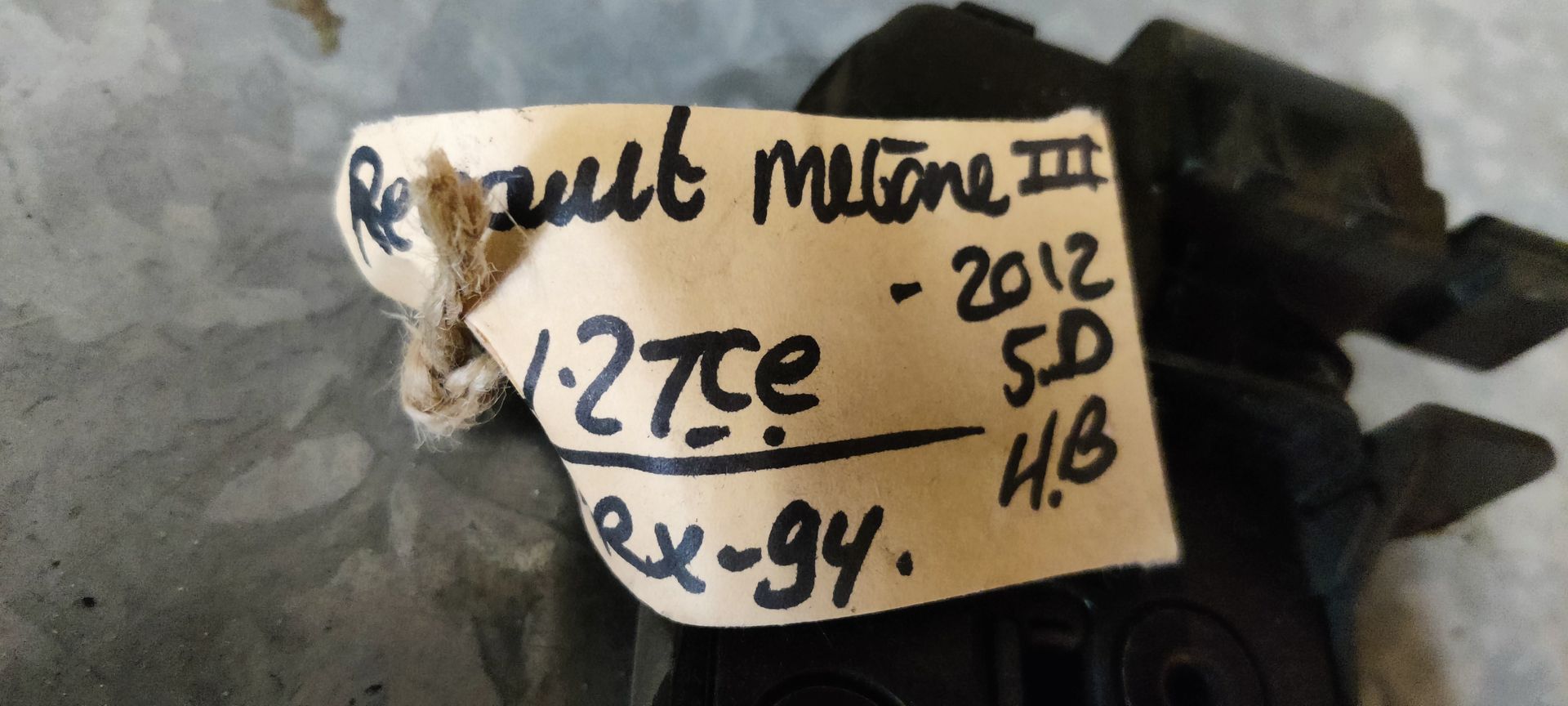 Een zwart metalen autodeurslot met een aangehecht identificatielabel, rustend op een grijze ondergrond.