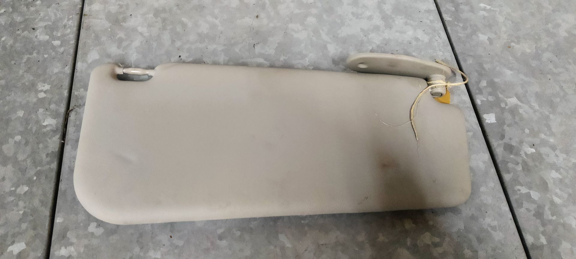 Een beige zonneklep voor de auto, inclusief bevestigingsbeugel en scharnier, ligt op een grijze tegelvloer.