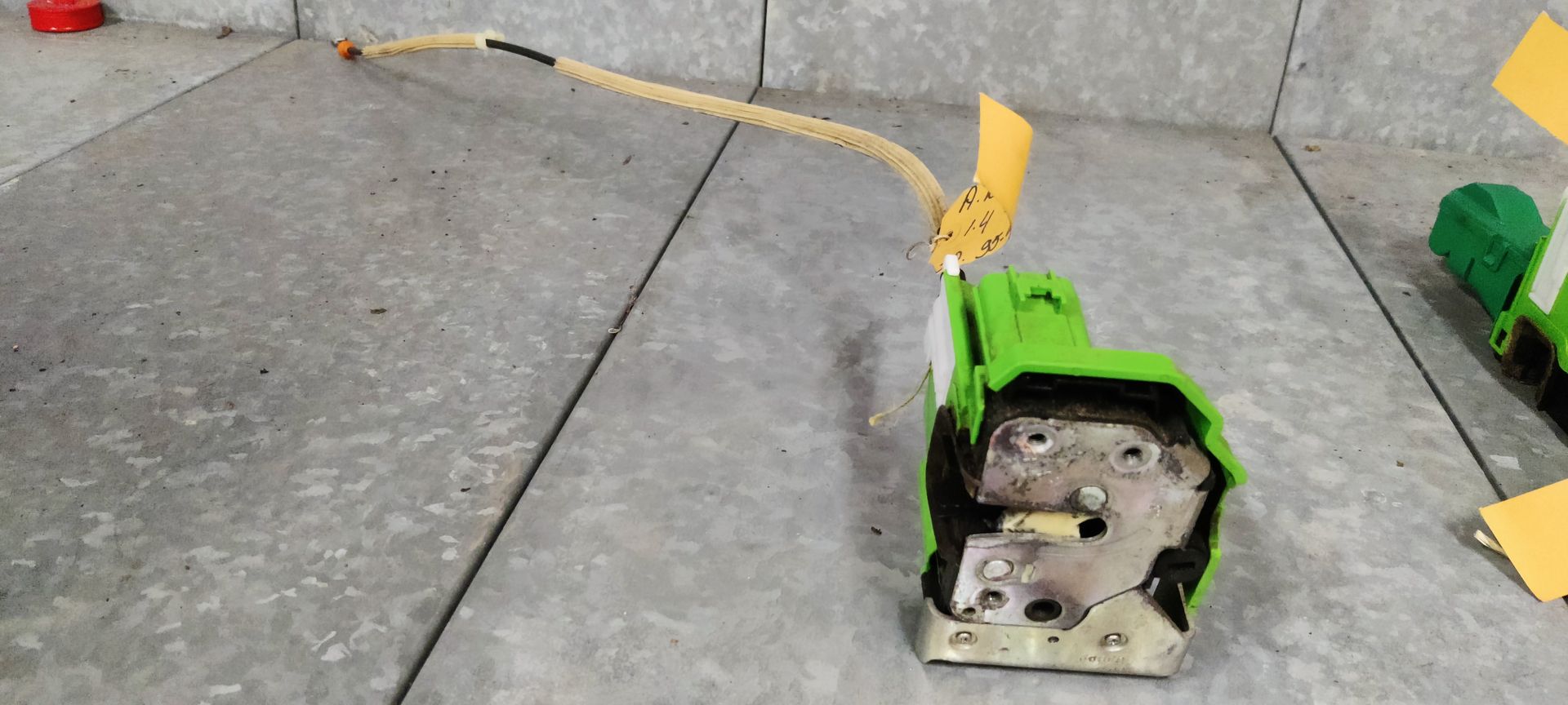 Een groen autodeurslotmechanisme met een aangesloten kabel, dat op een grijze tegelvloer ligt.