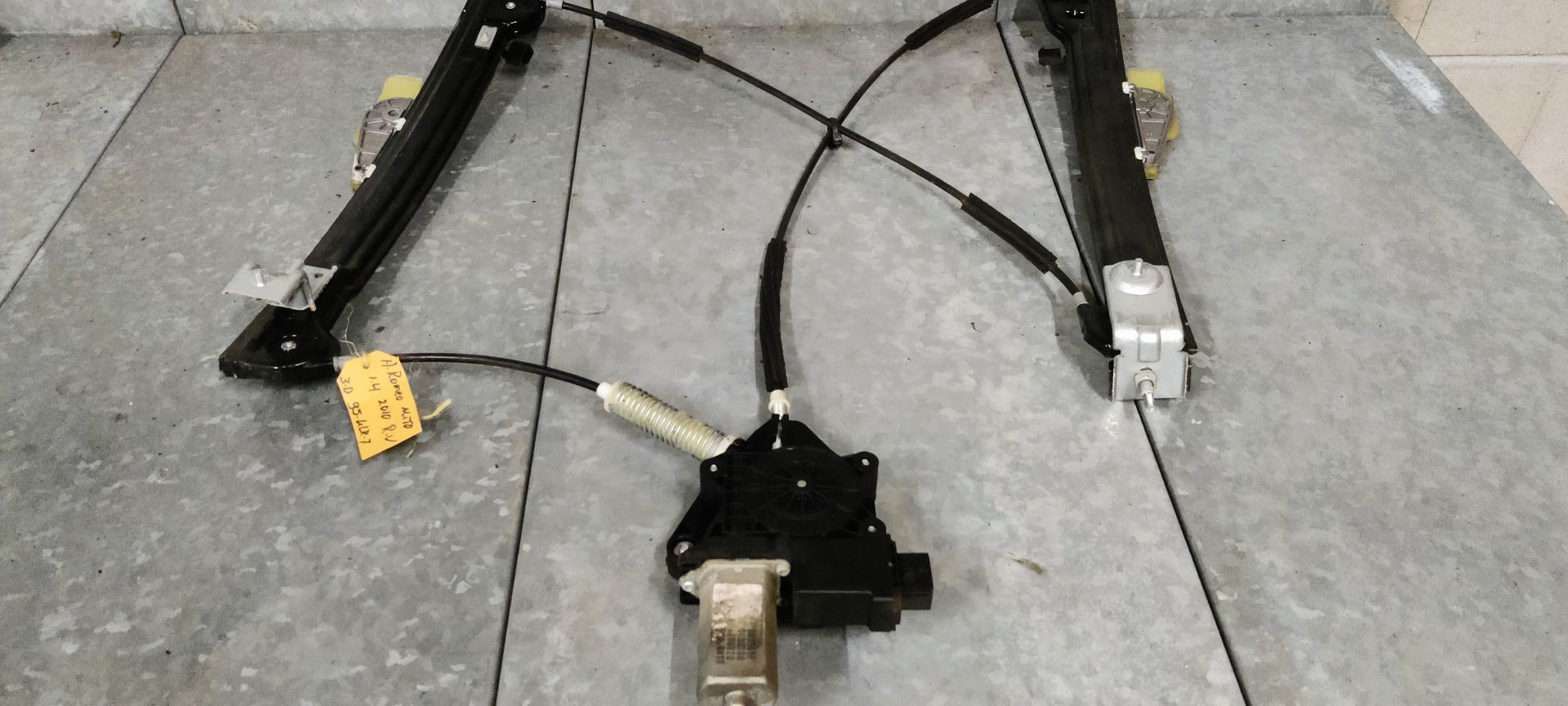 Een zwart raammechanisme voor een elektrische auto, inclusief motor en kabel, ligt uitgestald op een grijze vloer.