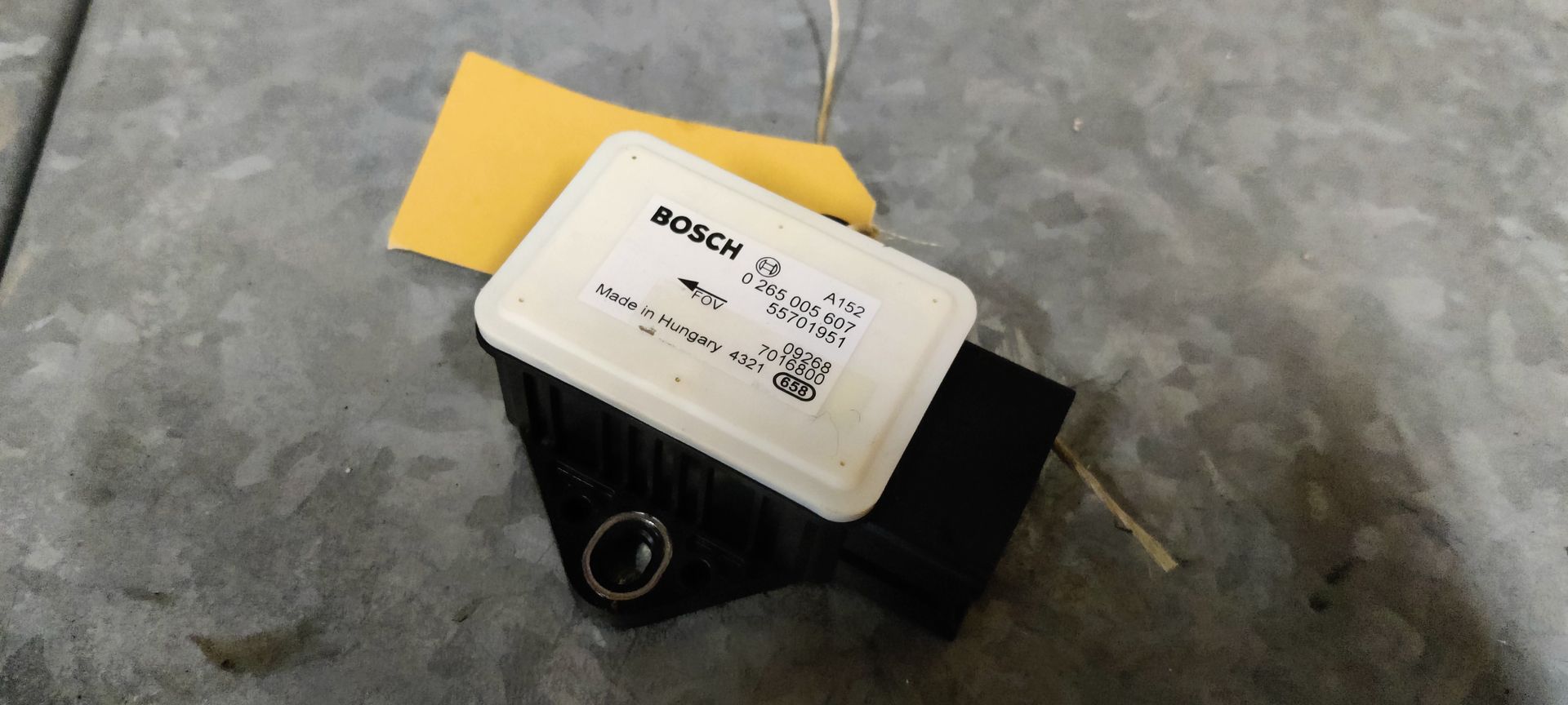 Een zwart-witte Bosch auto-elektronicaregeleenheid ligt op een grijs oppervlak met een geel label eraan.