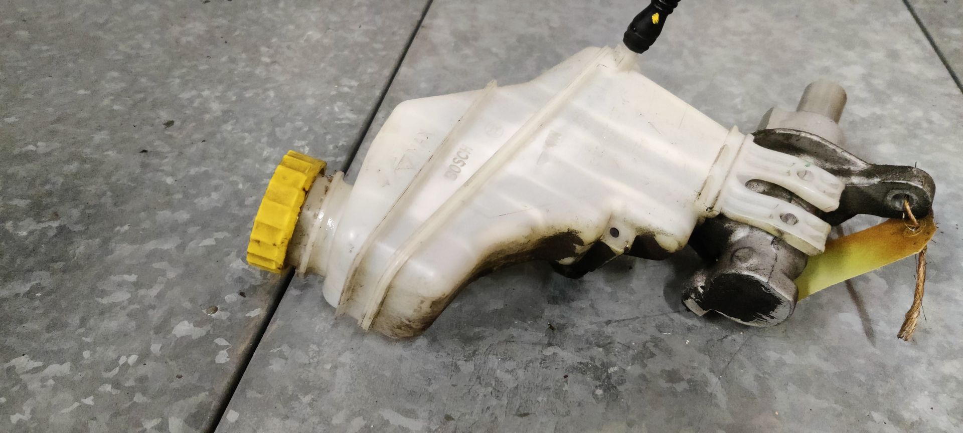 Een gebruikte hoofdremcilinder met een doorschijnend wit plastic reservoir en een gele dop staat op een grijze vloer.