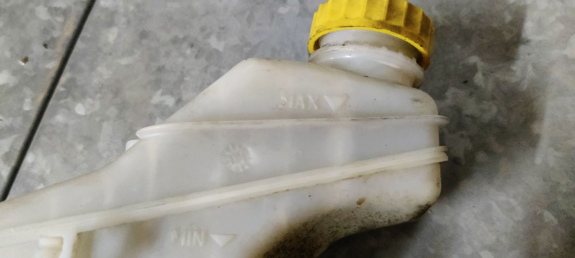 Een gebruikte hoofdremcilinder met een doorschijnend wit plastic reservoir en een gele dop staat op een grijze vloer.