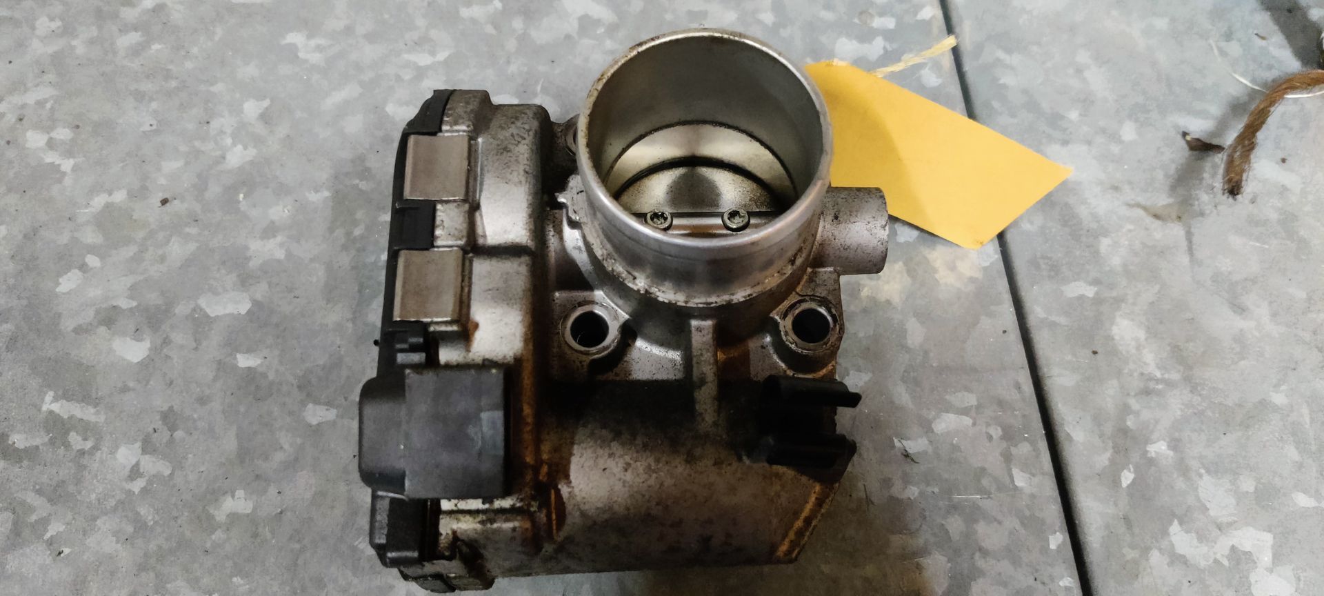 Een vuil, metalen gasklephuis van een auto ligt op een grijs, gestructureerd oppervlak met een geel label eraan.