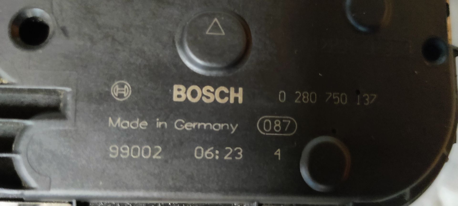 Een vuil, metalen gasklephuis van een auto ligt op een grijs, gestructureerd oppervlak met een geel label eraan.