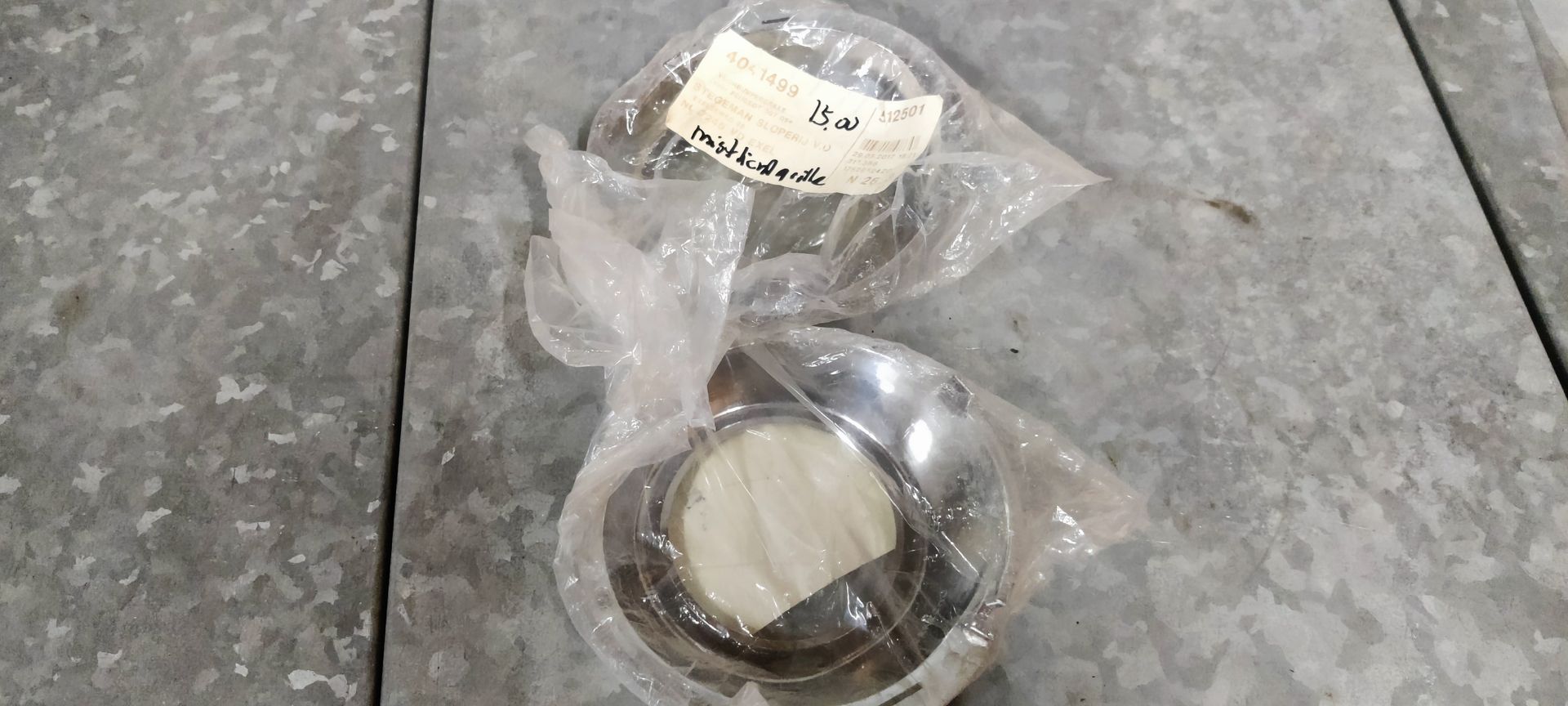 Twee chromen metalen wieldopringen in doorzichtige plastic zakjes, rustend op een grijs, gestructureerd metalen oppervlak.
