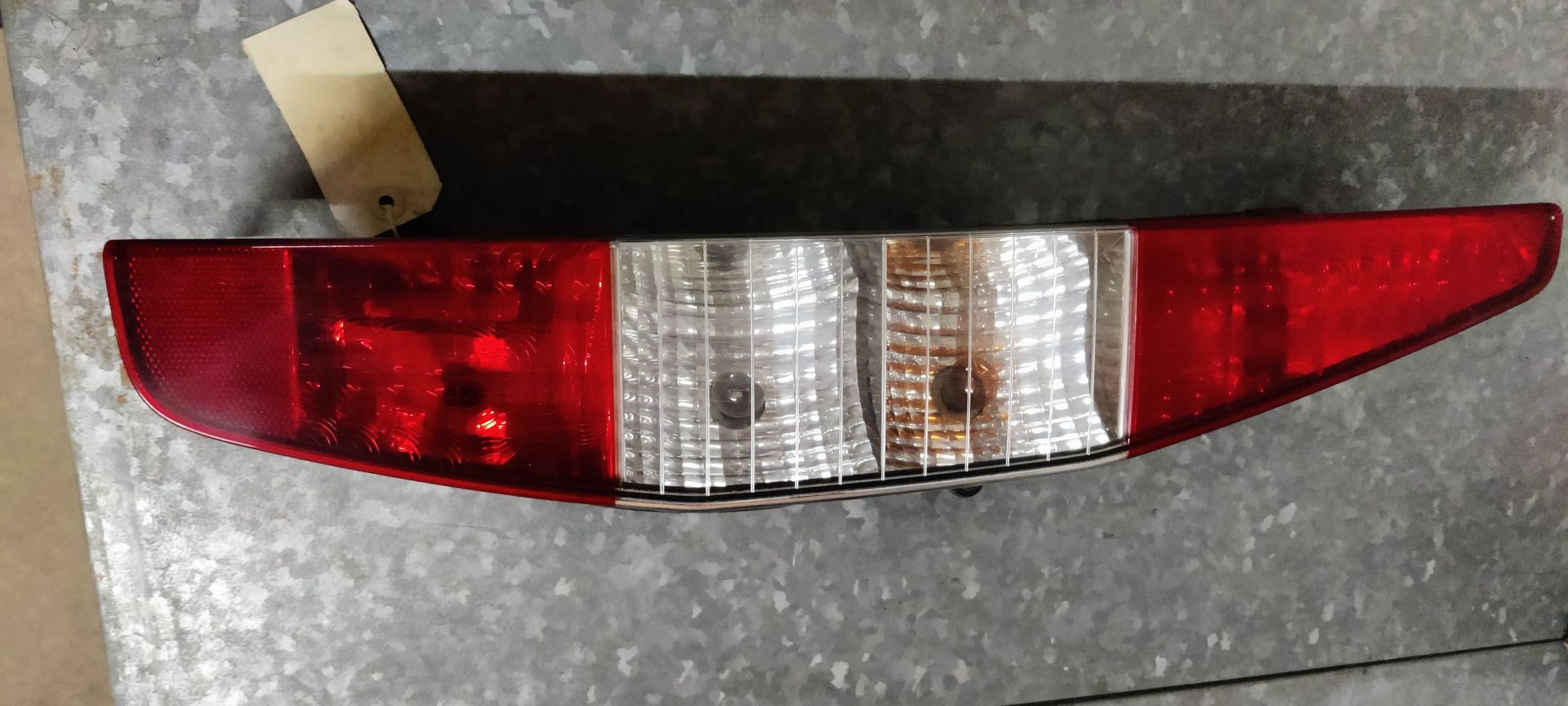 Een rode en transparante autoachterlichtunit met een klein labeltje eraan, rustend op een grijs metalen oppervlak.