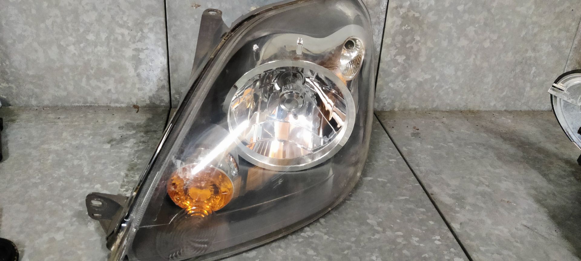 Een grijze autokoplampunit met een heldere hoofdlens en een amberkleurig knipperlicht, gemonteerd op een metalen oppervlak.