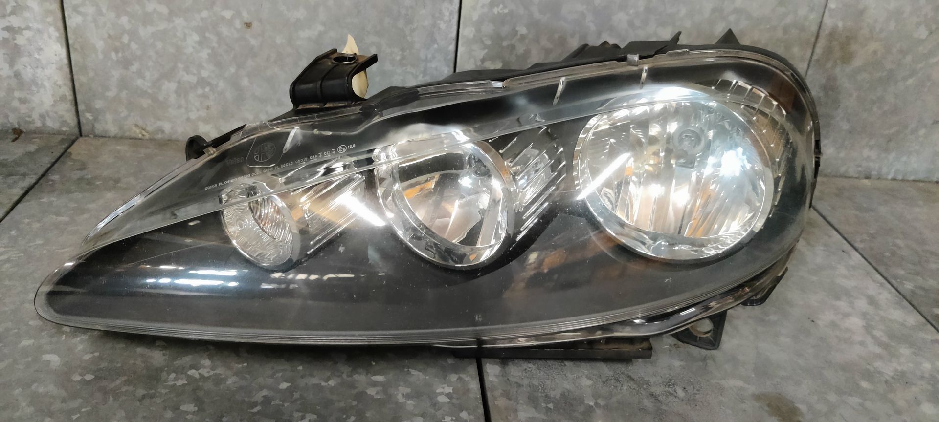 Een enkele autokoplampunit met een zwarte binnenbehuizing en drie ronde glazen lensdelen op een betonnen vloer.