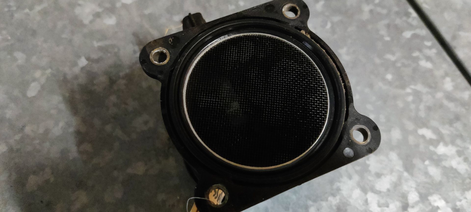 Een ronde, zwarte luchtmassameter voor auto's met een beschermend gaasfilter, van bovenaf gezien op een grijze ondergrond.