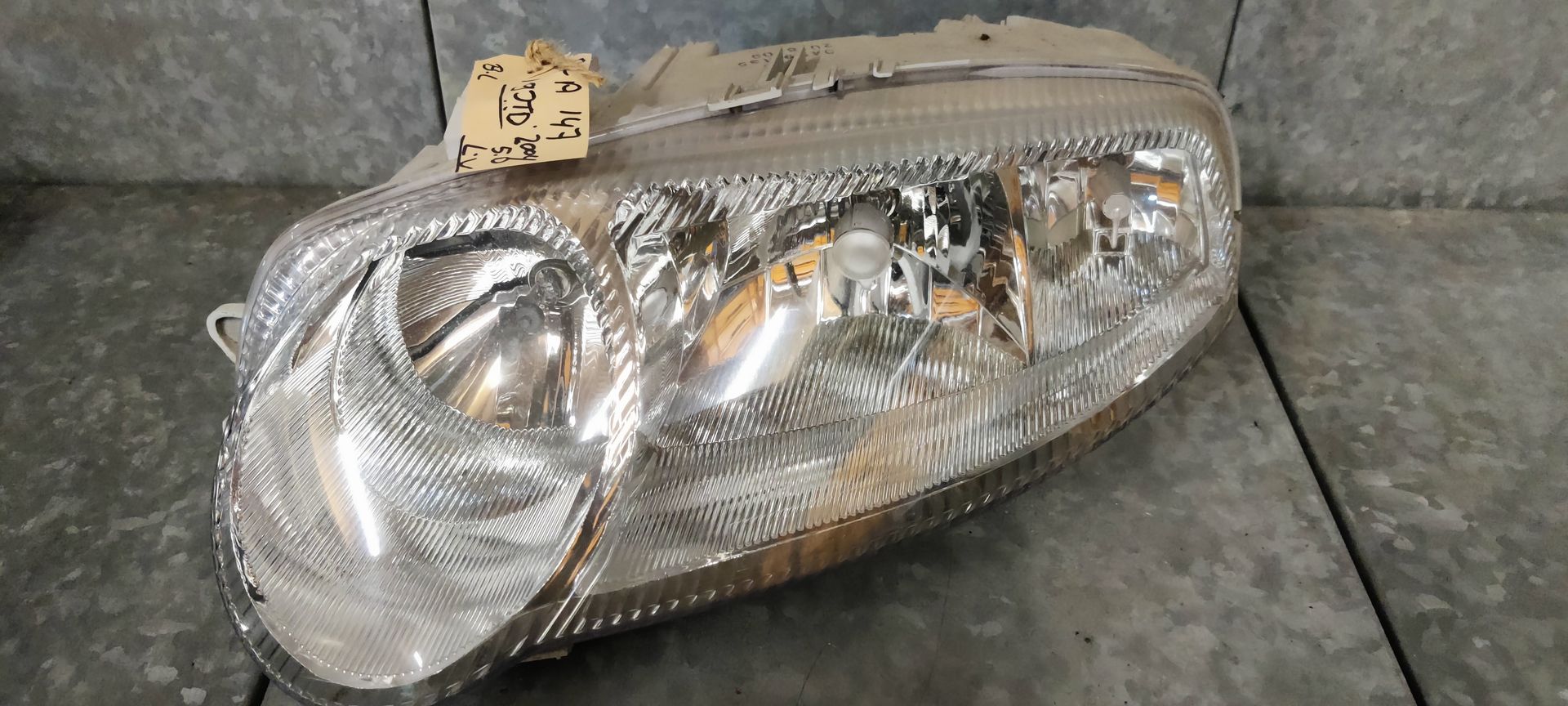 Een autokoplampunit met helder glas, verchroomde binnenreflectoren en een aangehecht identificatielabel.